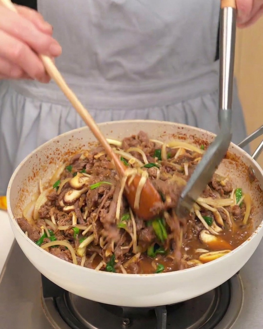 調理された牛肉、きのこ、玉ねぎ、ねぎを木製のスプーンとトングで混ぜ合わせている様子。