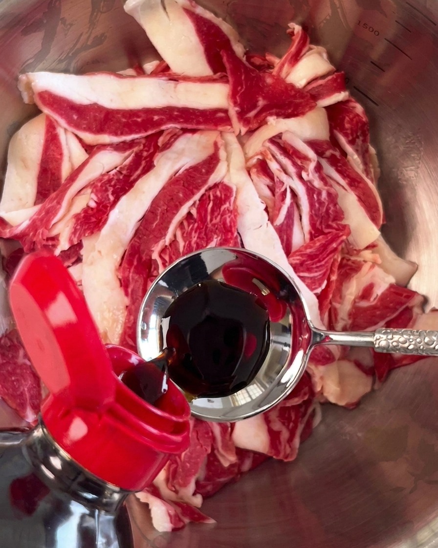 ボウルに入った牛肉に、金属製のスプーンから濃口醤油を注いでいる様子。
