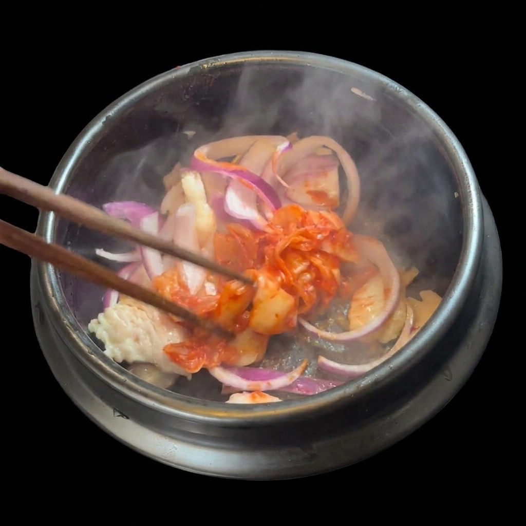 石鍋の中の豚肉と玉ねぎに、刻んだ真っ赤なキムチを加えている様子。