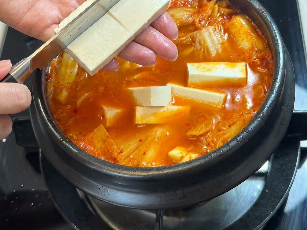 グツグツ煮えるピリ辛のキムチチゲの上で、豆腐を直接包丁でカットして入れている様子。