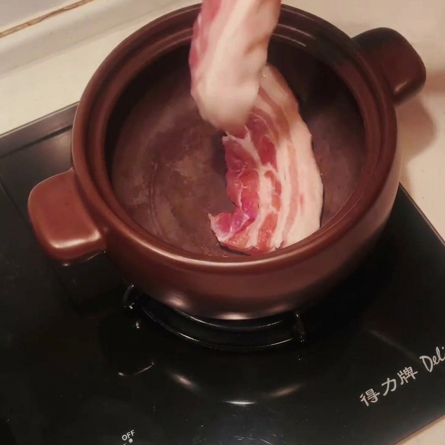 予熱した黒い土鍋にトングで生の豚バラ肉を入れている様子。