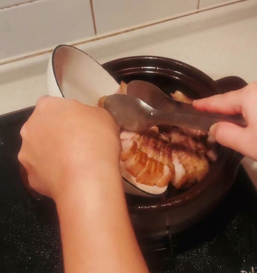 焼いた豚バラ肉の切り身を白いボウルから黒い土鍋に戻し入れている様子。