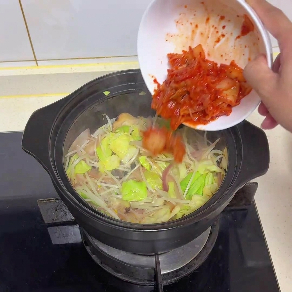 野菜が炒められた鍋に、真っ赤な刻みキムチを入れている様子。