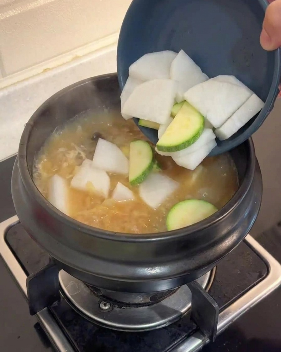 煮立っている茶色のスープが入った鍋に、青いボウルから大根とズッキーニを入れている様子