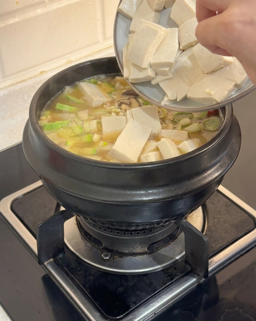 湯気の立つ韓国チゲの鍋に、手を使って四角い豆腐をそっと入れている様子