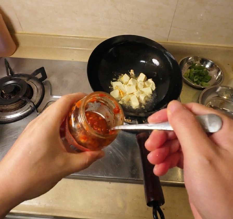 豆腐が入った中華鍋に、スプーンで真っ赤なチリソースを加えている様子。