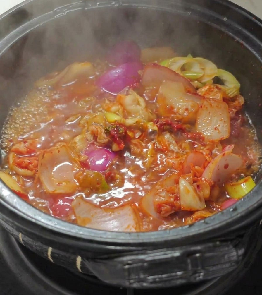 炒めたキムチと玉ねぎが入った黒い石鍋に熱湯を注ぎ、真っ赤なスープを作っている様子。