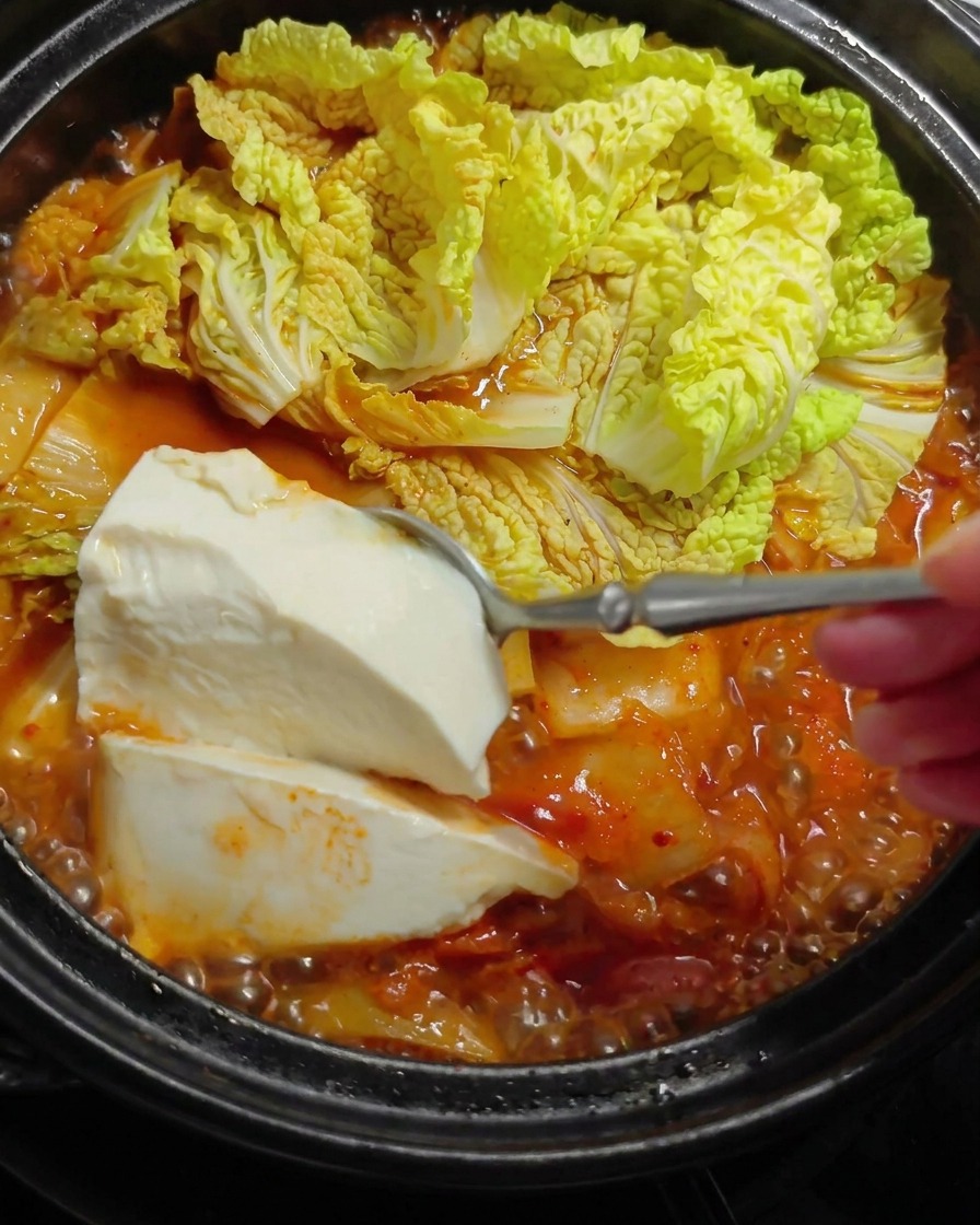 大きなスプーンで柔らかい白い豆腐をすくい、白菜が入った熱々の赤いスープの中にそっと入れている様子。