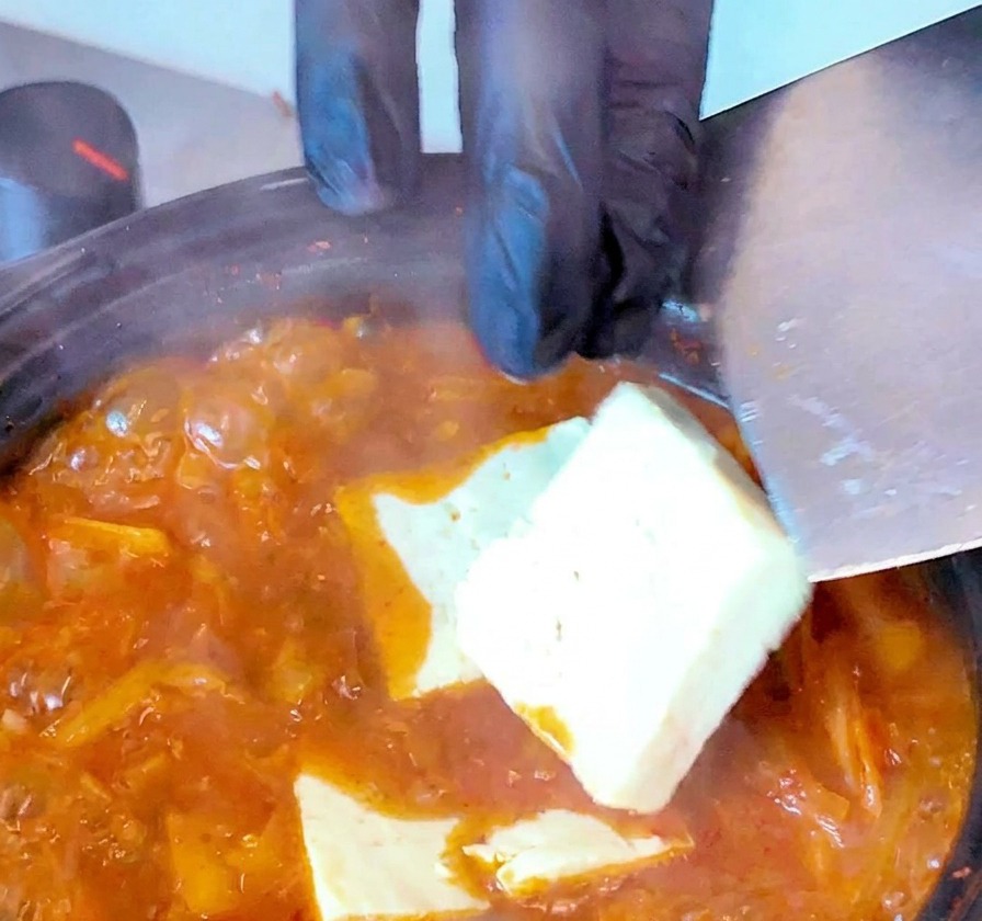 ぐつぐつと煮える赤いキムチスープに、ヘラを使って白い豆腐を並べて入れている様子。