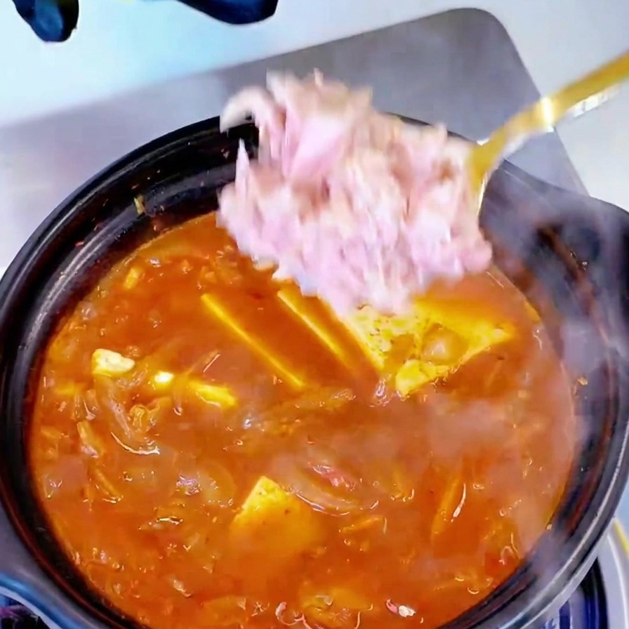 沸騰しているキムチと豆腐のスープに、スプーンでツナを塊のまま加えている様子。