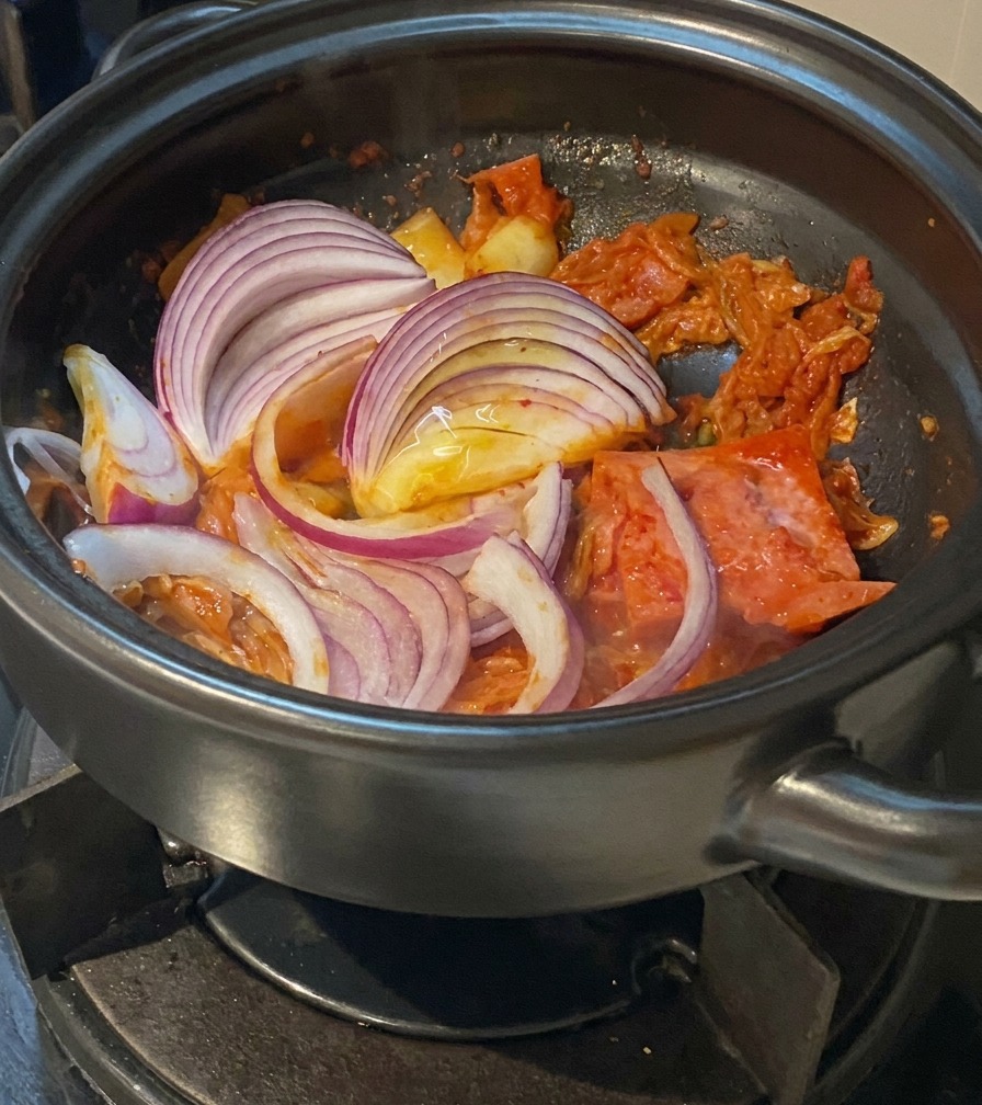 キムチと豚肉が入った鍋に、スライスした赤玉ねぎを加えて混ぜている様子。