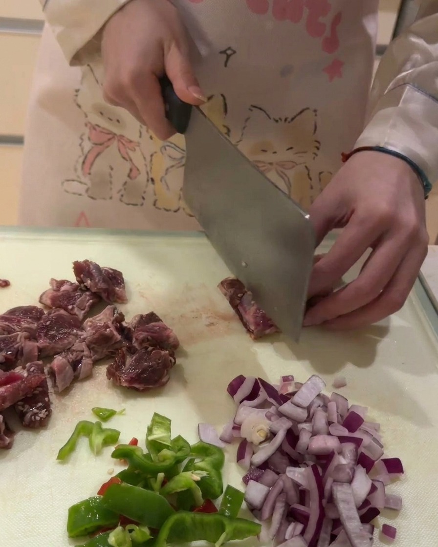 白いまな板の上で、生の牛バラ肉を一口大の角切りにしている様子。