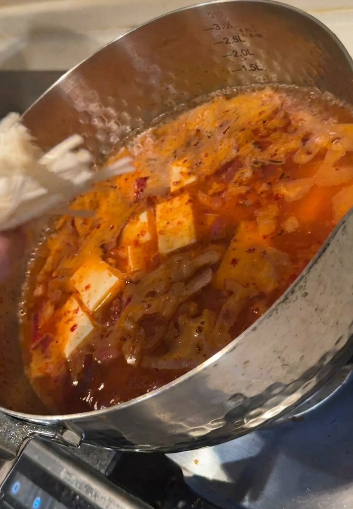 豆腐の角切りが入った赤いスープに、えのき茸を一束加えている様子。