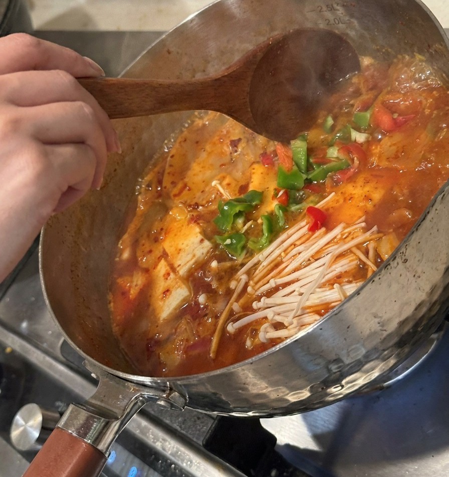 煮立っている鍋に、刻んだピーマンとキムチを加え、木べらで混ぜている様子。