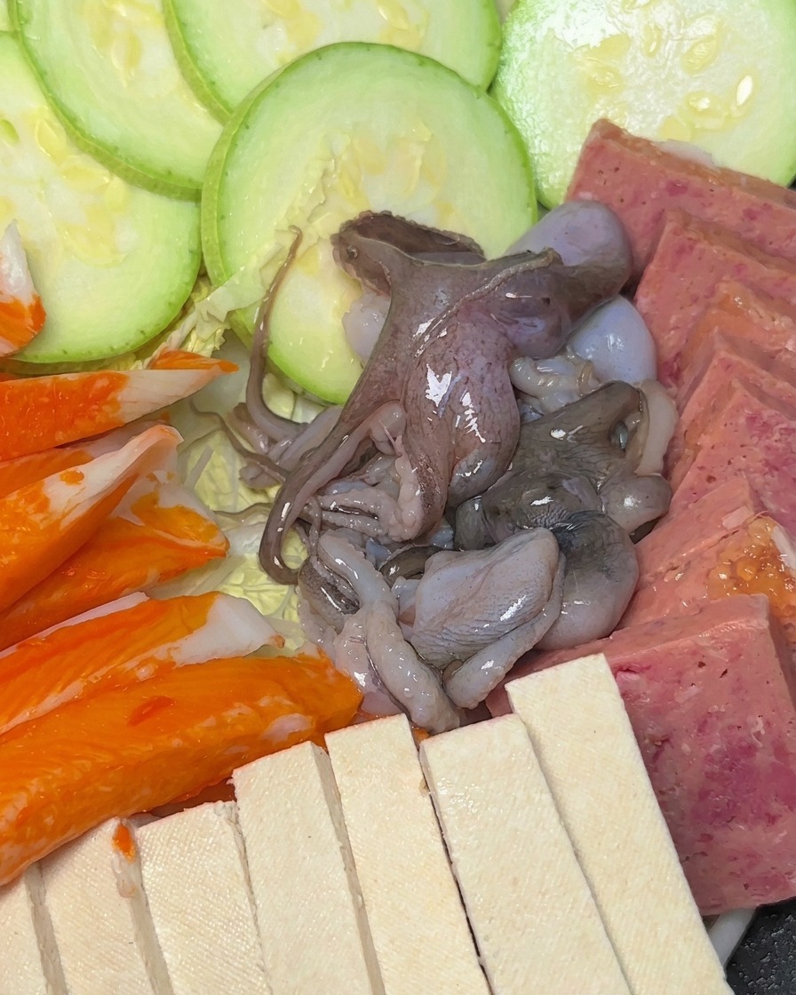 ズッキーニやスパムが並んだ鍋の中に、カニカマ、タコ、しいたけを配置した様子