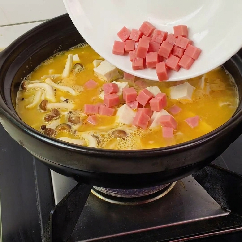 黄金のスープに、さいの目切りのハム、豆腐、きのこ、グリーンピースを加えている様子。