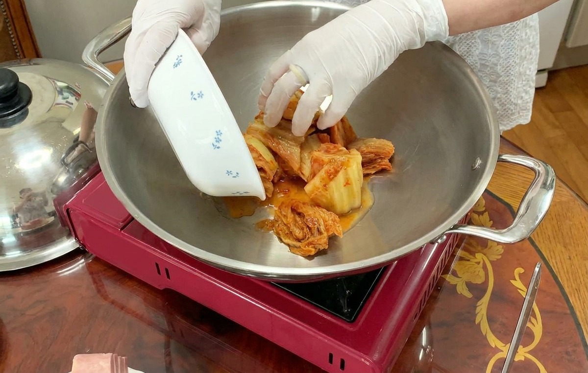 大きな銀色の鍋の中央に、小さな器からカットされたキムチを移している様子。