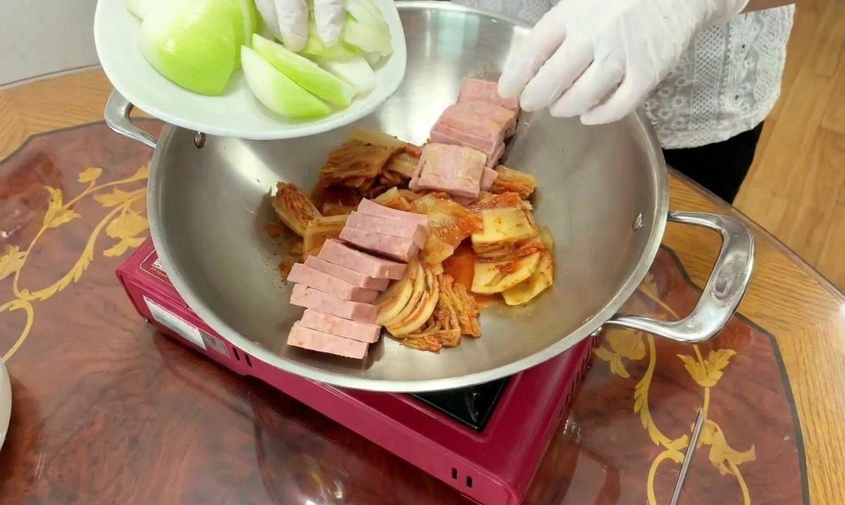 キムチとスパムが入った鍋に、くし形に切った白い玉ねぎを加えている様子。