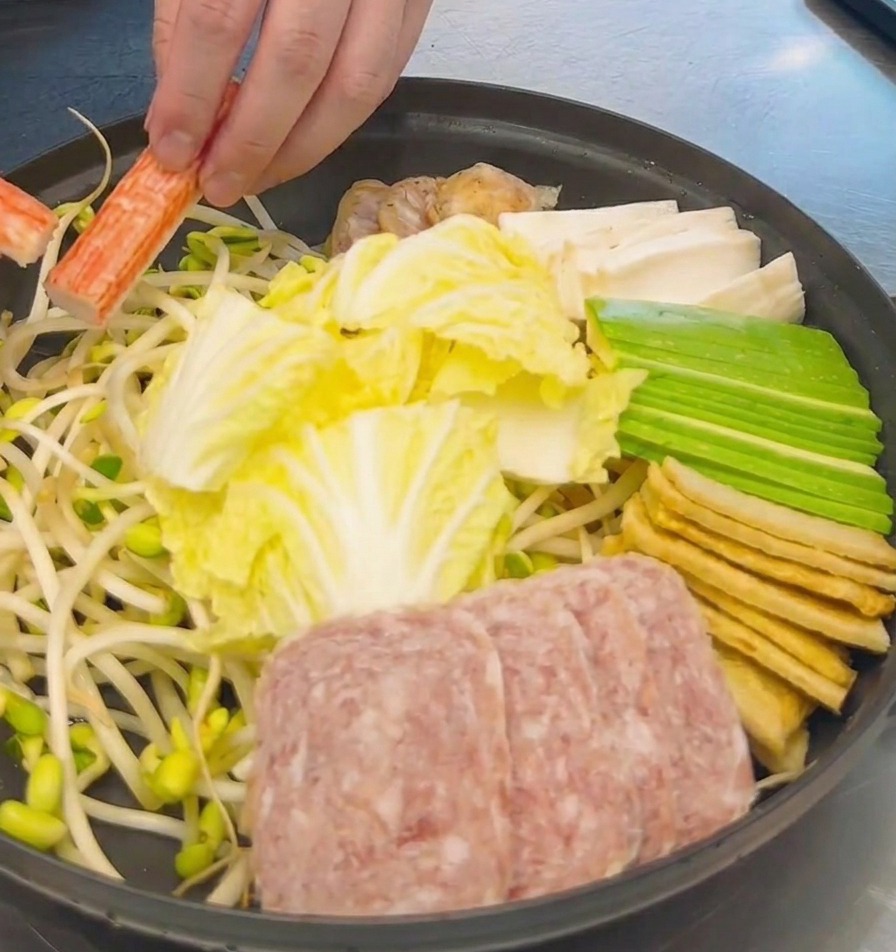 白菜、スパム、豆腐が並んだ鍋に、オレンジと白のカニカマを盛り付ける様子。