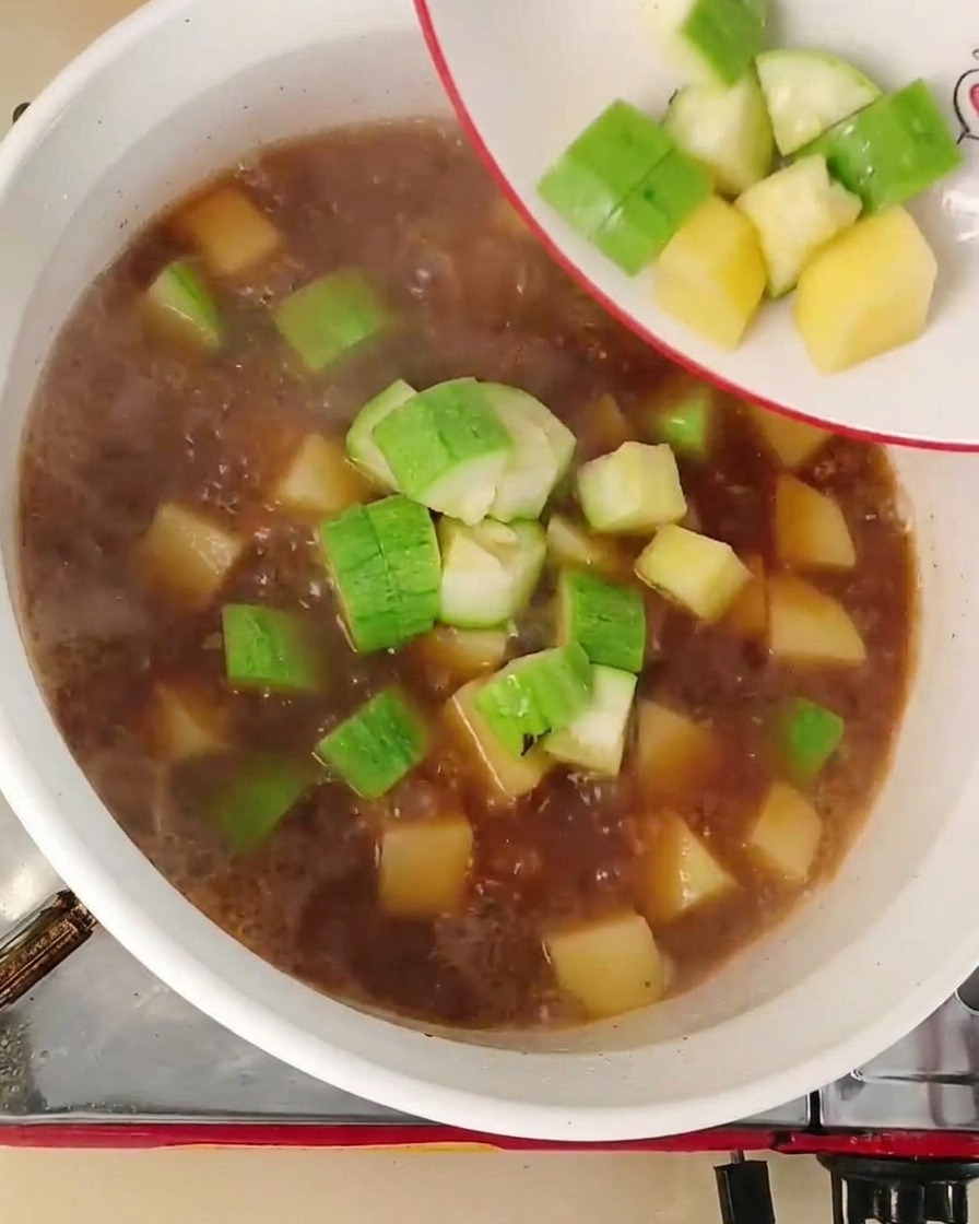 角切りにした緑のズッキーニと黄色い玉ねぎを、小さな白いお皿から赤茶色の煮え立つ鍋のスープに流し入れている様子。