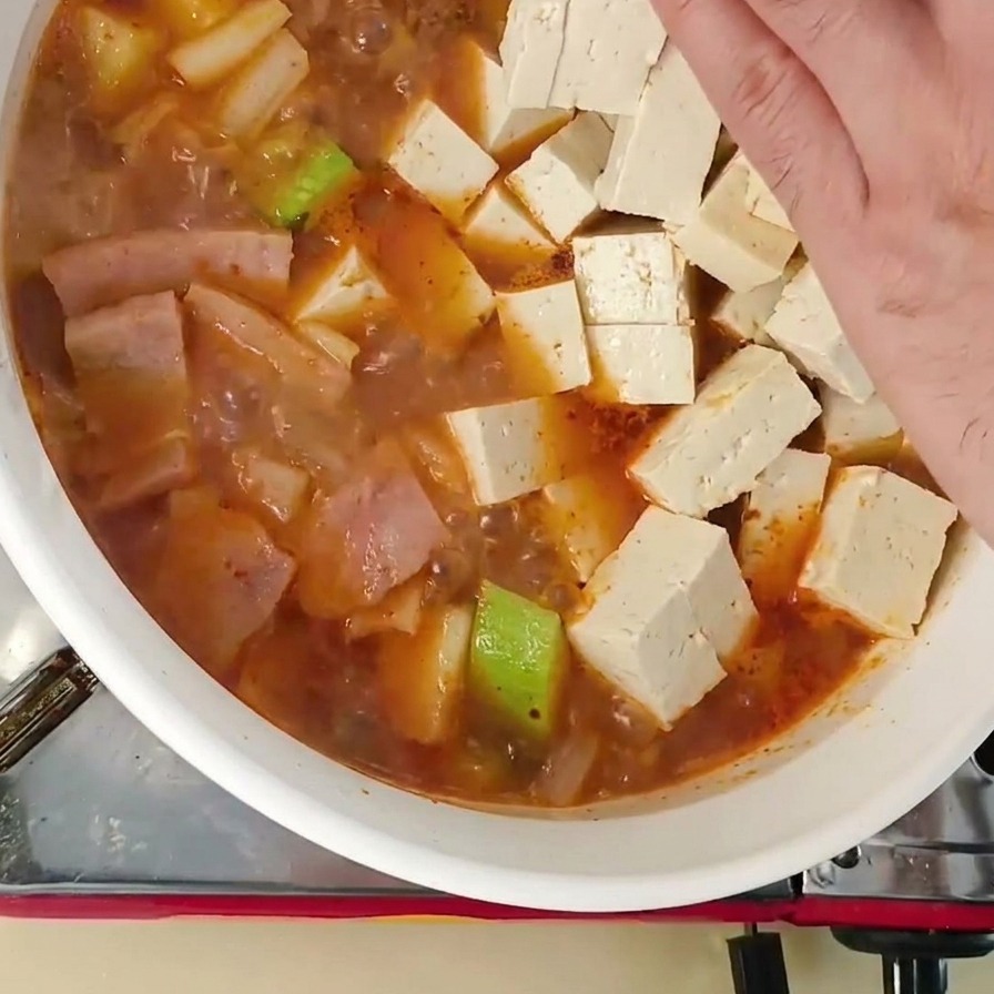 ズッキーニなどの具材が入った韓国味噌チゲの煮え立つ鍋に、新鮮な白い豆腐の角切りをそっと手で入れている様子。