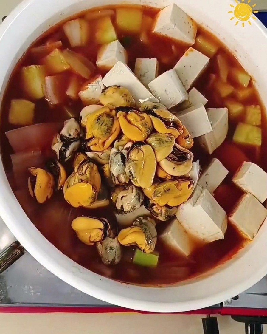 豆腐や野菜がたっぷり入った沸騰した鍋の中央に、オレンジ色のムール貝の身と、大きくグレーがかった生の大きなエビを加えている様子。