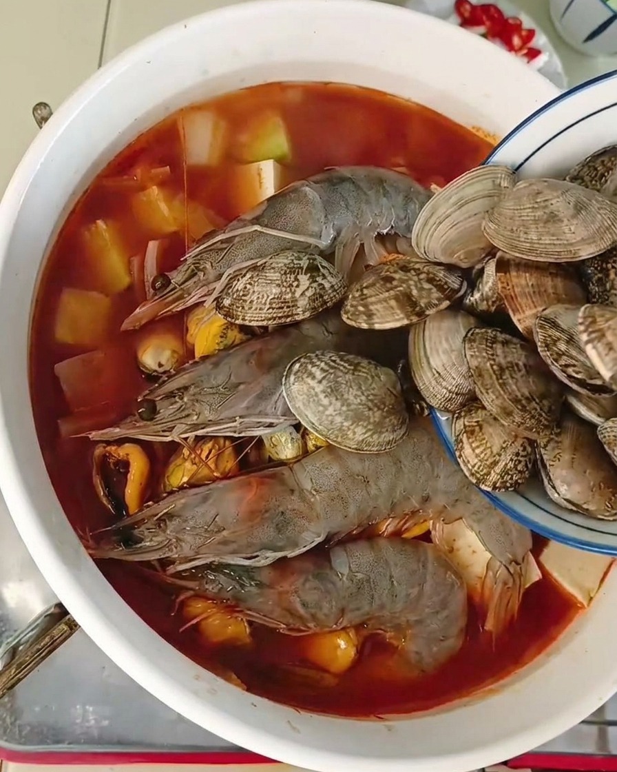 青い縁取りの小さなボウルから、エビ、ムール貝、豆腐がすでに入っている赤いスープの鍋に、新鮮な殻付きあさりを流し入れている様子。