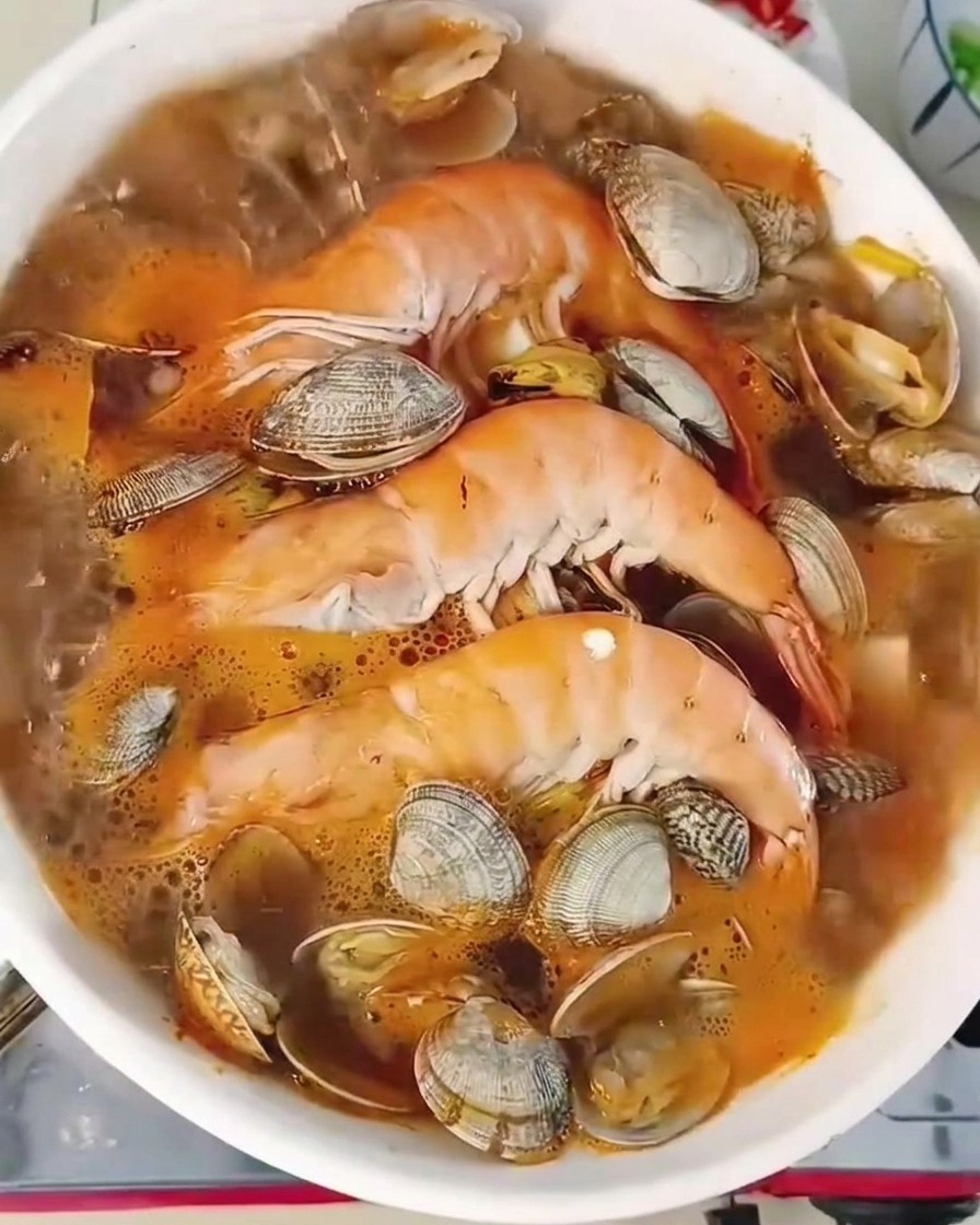 エビ、あさり、豆腐が入った白い鍋の中でふつふつと煮え立つ海鮮チゲのクローズアップ。表面には新鮮な青唐辛子と長ねぎが散らされている。