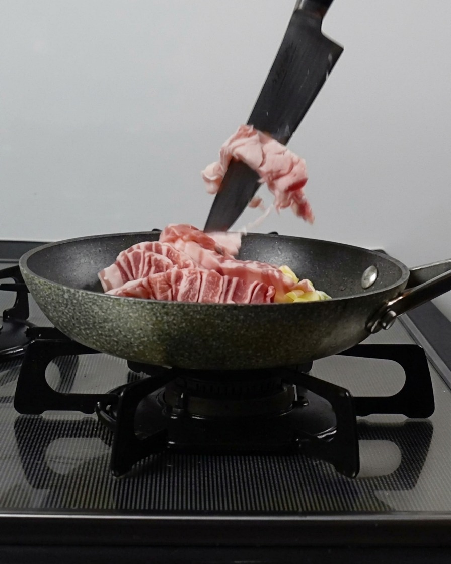 包丁を使って、豚バラ肉、ねぎ、にんにくを熱したフライパンに滑り込ませる様子。