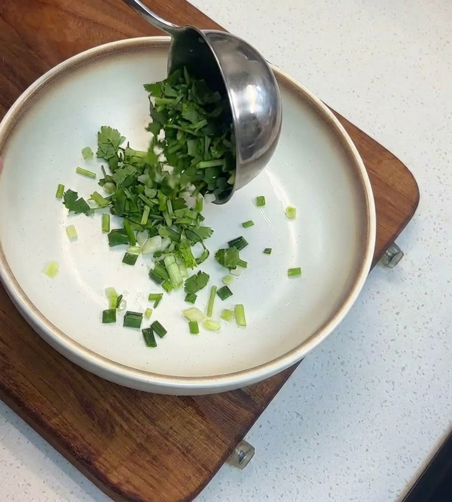 縁が茶色の白いボウルに、お玉でたっぷりの刻んだパクチーとねぎを入れている様子。
