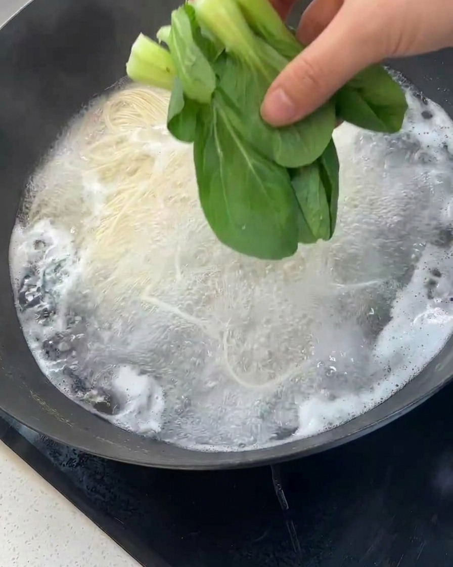 麺が茹でられている中華鍋の中に、手で新鮮な青梗菜を投入している様子。