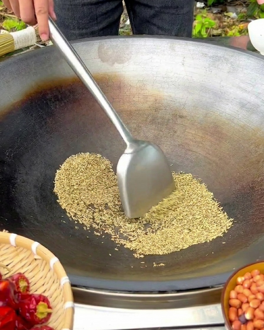 弱火にかけた使い込まれた大きな鉄鍋で、金属製のヘラを使って材料を炒めている様子。