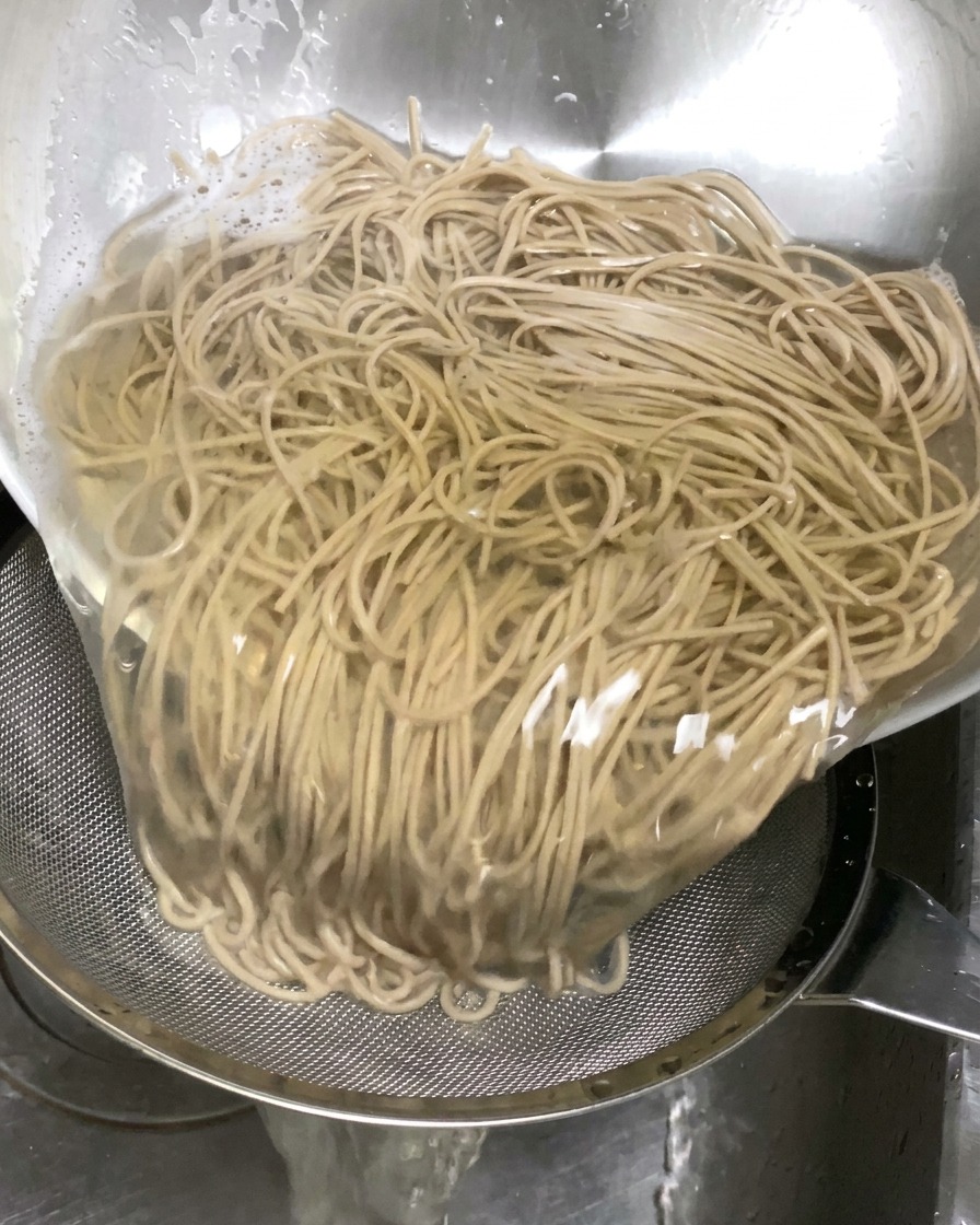 ボウルに入れた茹でたてのそばを、シンクの中でザルにあけて湯切りしている様子。