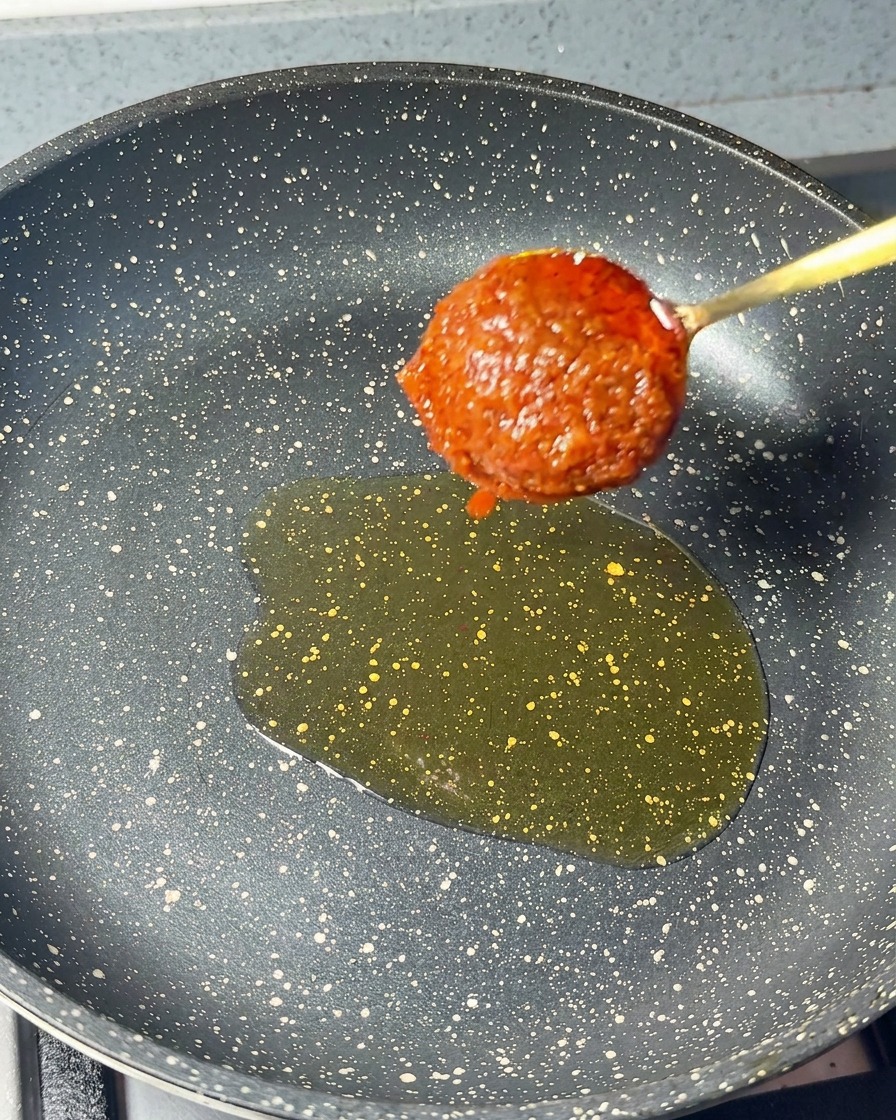 温まったごま油に、濃厚な赤色のエビペーストをスプーンで加えている場面。