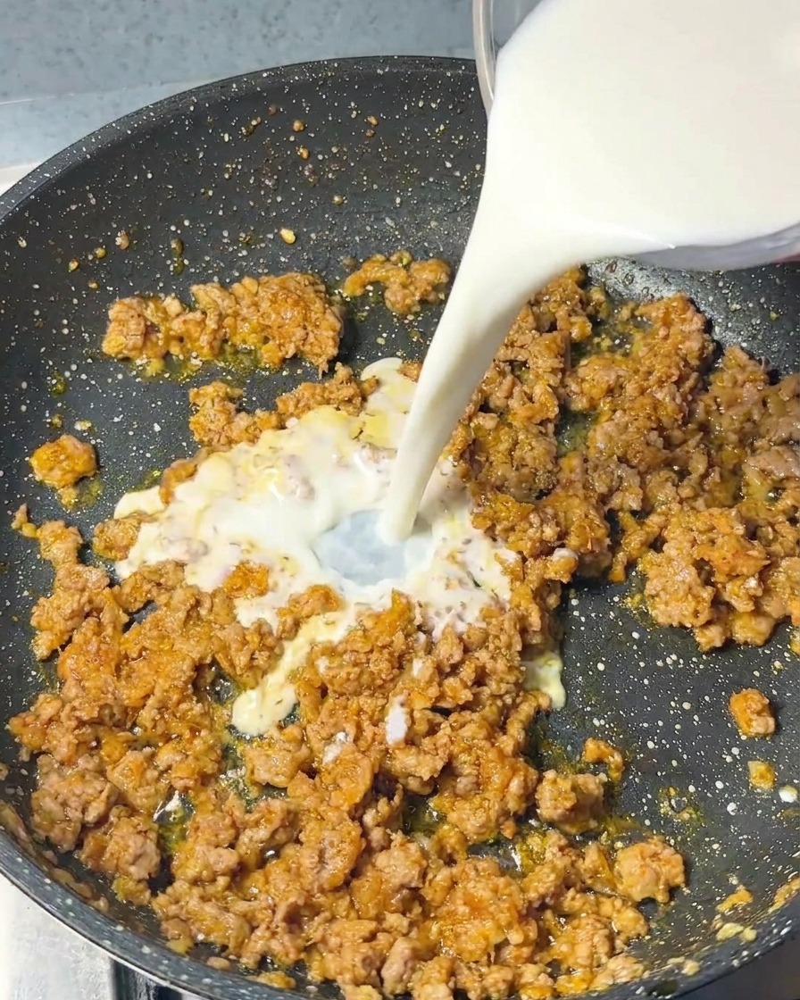 炒めたひき肉の入ったフライパンに、白い牛乳をゆっくりと注ぎ入れている様子。