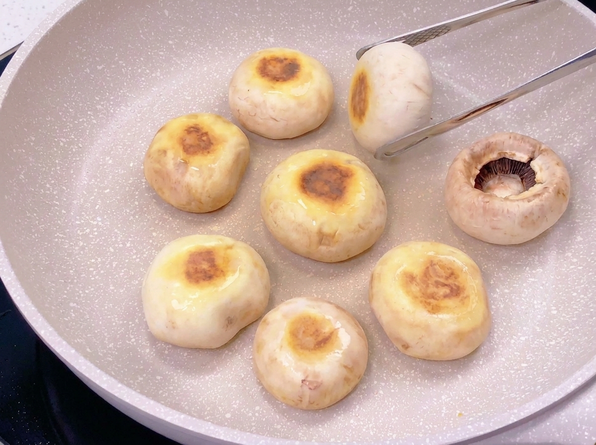 フライパンに並んだマッシュルームの傘を裏返し、表面についた丸く茶色い焼き色を見せている様子。