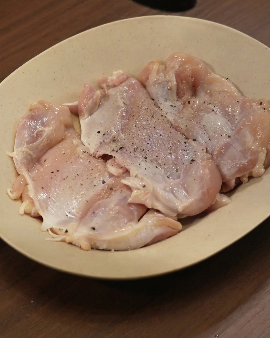 ベージュの皿に並べた鶏もも肉に黒こしょうや各種スパイスを振りかけている様子