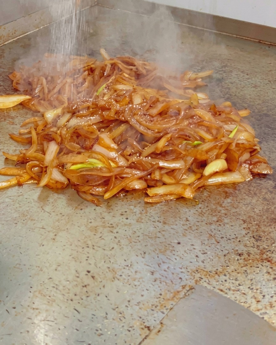 熱い鉄板の上で、湯気を立てる艶やかなあめ色玉ねぎの山に、微粒塩が振りかけられている様子