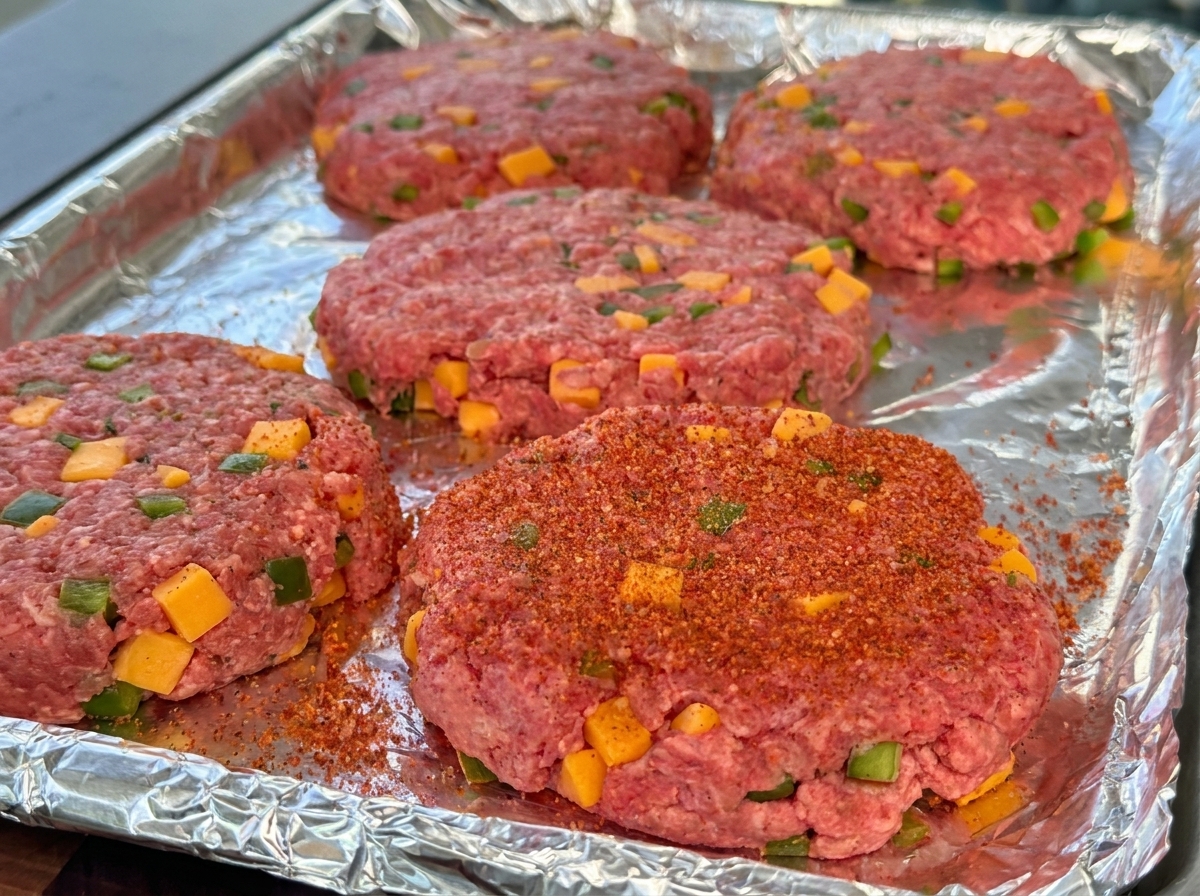 アルミホイルを敷いたトレイの上に並べられた、チーズとピーマンが混ざった生のビーフパティ。赤いBBQパウダーで味付けされている。