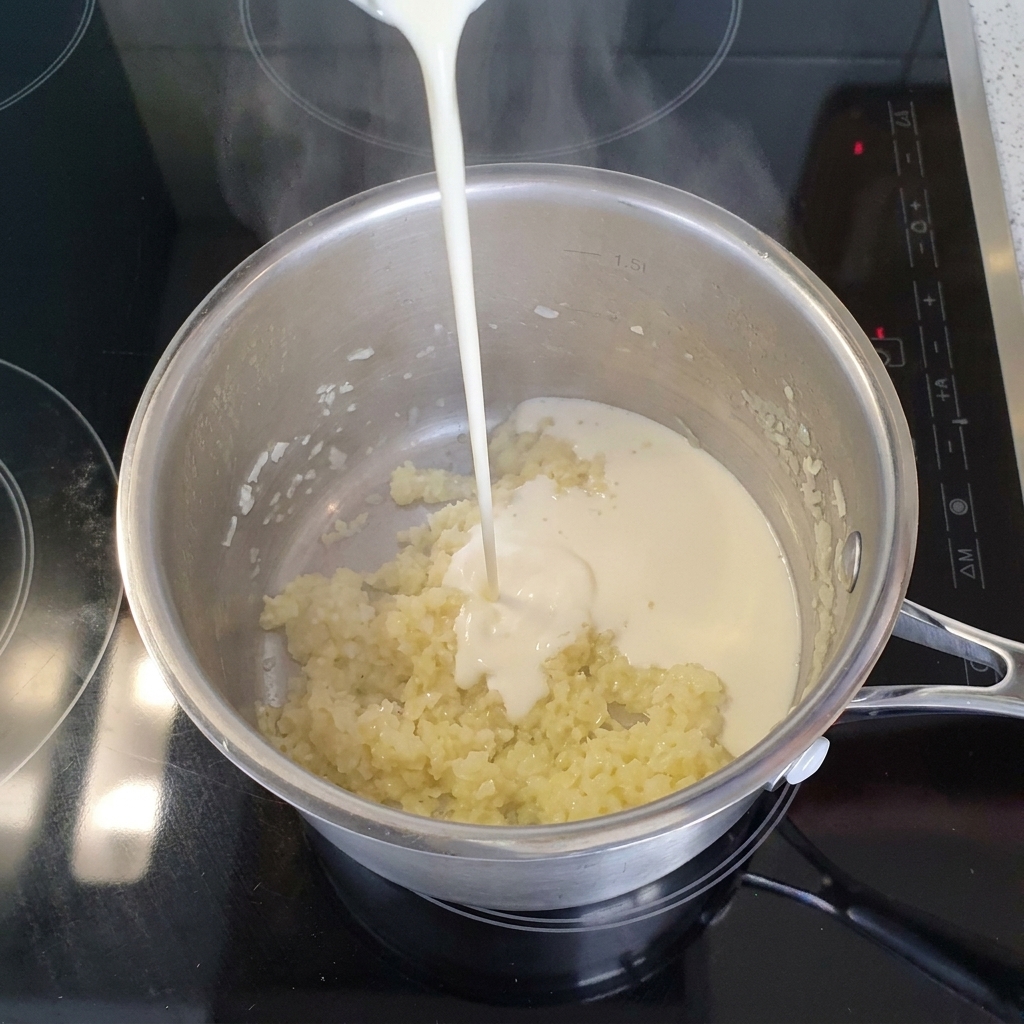 みじん切りの玉ねぎとニンニクが炒まったステンレスの鍋に、さらっとしたライトクリームが注がれている様子。