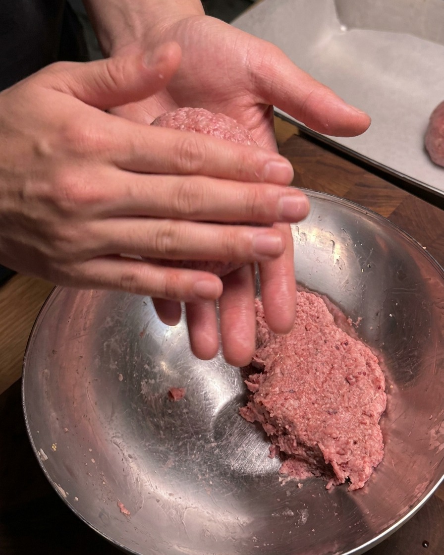 下味をつけた生の牛挽き肉を、両手でしっかりと丸めてきれいな球体の肉団子に成形する手元のアップ。