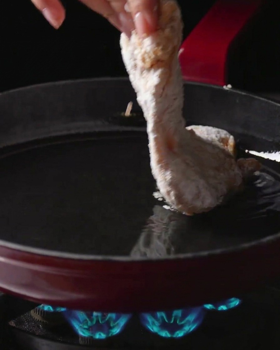火のついたガスコンロの上の熱い油が入ったフライパンに、衣をつけた鶏もも肉を手で丁寧に入れている様子。