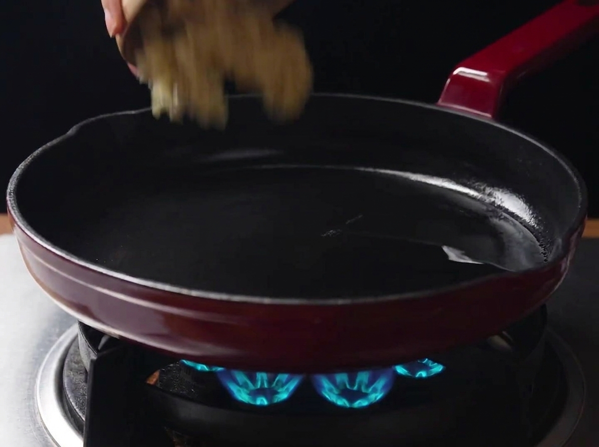 火のついたガスコンロの上で、鶏肉を炒めた後の旨味が残る熱いフライパンに、小さなボウルから刻んだにんにくを加えている様子。