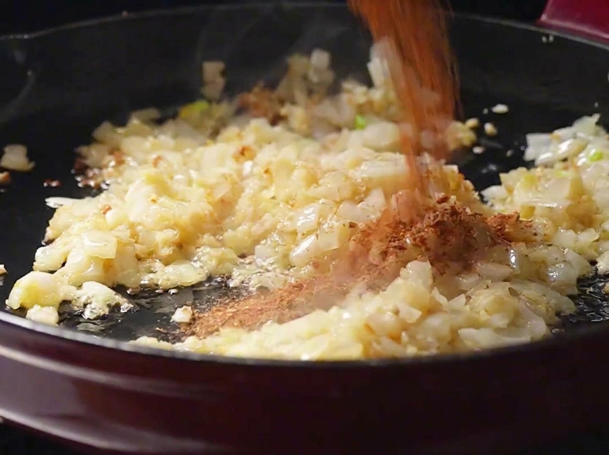 フライパンの中で炒められた玉ねぎとにんにくの上に、自家製の赤茶色のケイジャンスパイスパウダーを振りかけている様子。
