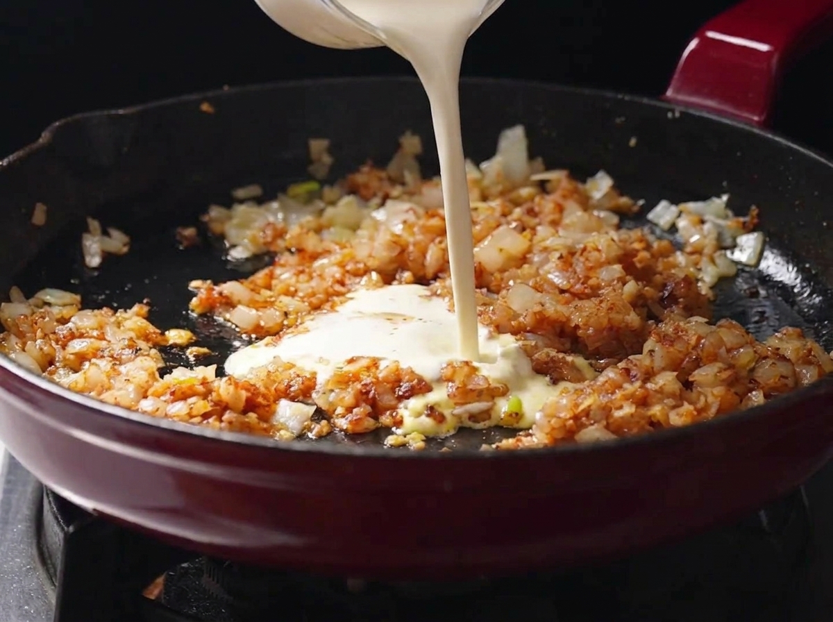 スパイスをなじませた香味野菜が入ったフライパンに、ガラスの容器から生クリームを注ぎ入れている様子。