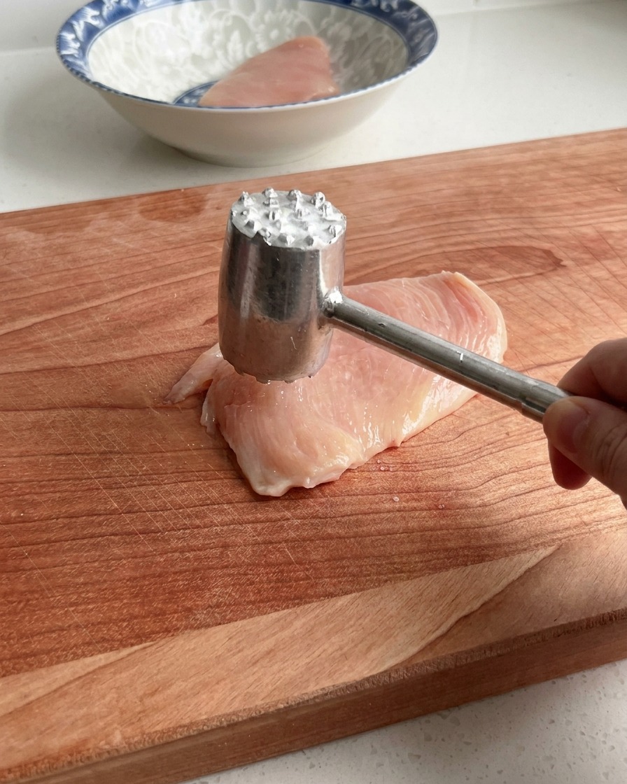 大きな木製のまな板の上で、ミートハンマーを使って生の鶏むね肉を優しく叩く様子。