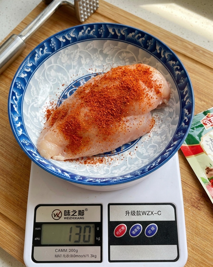 デジタルキッチンスケールの上に置かれた陶器のボウルの中で、オレンジ赤色のシーズニングパウダーをたっぷりとまぶされた生の鶏むね肉。