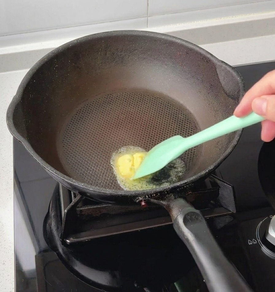熱した黒いフライパンの中央で、緑色のシリコンスパチュラを使って小さな角バターを溶かしている様子。