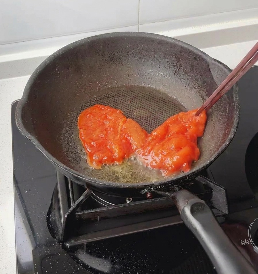 箸を使って、赤くしっかりと漬け込まれた鶏むね肉を、軽く油を引いた黒いフライパンに入れる様子。