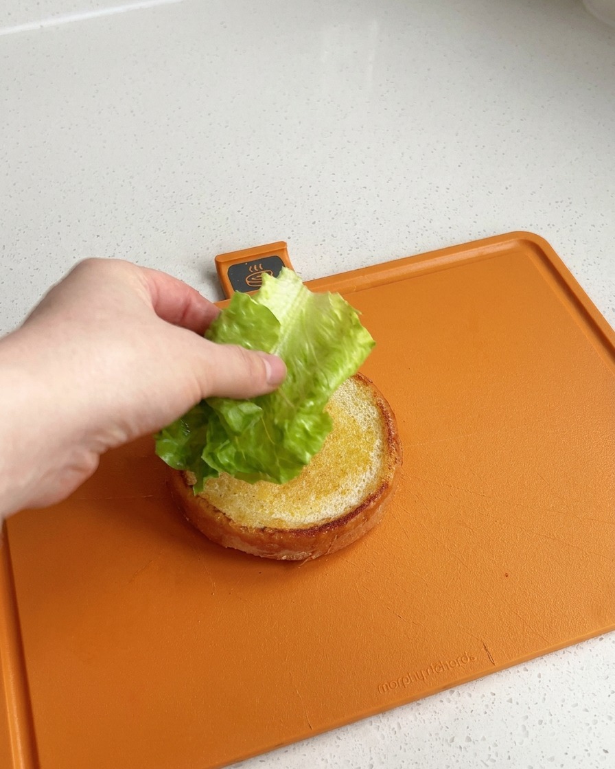 オレンジ色のまな板の上で、きれいにトーストされたバーガーバンズの下半分に、新鮮な緑のレタスを乗せている手。